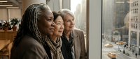 Diverse group of three professional women standing together at office window overlooking urban cityscape, representing corporate teamwork, leadership, and business collaboration in modern workplace setting.