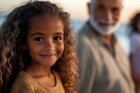 Young African American girl with curly hair smiling warmly at camera, wearing yellow sweater, with senior man in background during golden hour lighting.