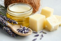 Natural cosmetic ingredients with coconut oil in glass jar, shea butter cubes, dried lavender flowers on wooden spoon against light background.
