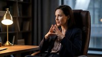 Professional businesswoman in dark suit sitting thoughtfully in modern office with warm lighting and city view background for corporate presentations.