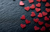 Red glitter heart confetti scattered on dark slate background, creating romantic Valentine's Day or wedding celebration atmosphere.