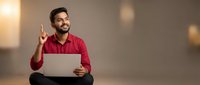 Young Indian businessman in red shirt pointing up with laptop showing eureka moment during creative brainstorming session in modern office space.