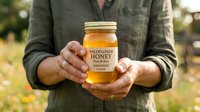 Beekeeper holding jar of wildflower honey outdoors in natural light, showcasing raw organic honey production and sustainable farming practices.