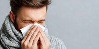 Caucasian man with gray scarf blowing nose into tissue, eyes closed with discomfort, showing cold or flu symptoms against neutral background.