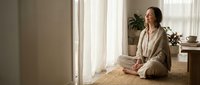 Young Caucasian woman in beige robe practicing mindful meditation sitting cross-legged on wicker mat by bright window in minimalist home interior.