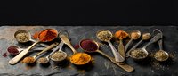 Colorful spices and herbs arranged in wooden spoons and bowls on dark slate background for cooking and culinary seasoning applications.