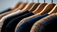 Warm knitted sweaters in beige, black and navy blue colors hanging on wooden hangers in closet, showing texture and fabric details. Cozy winter wardrobe.