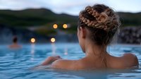 Young woman with braided updo relaxing in infinity pool at dusk, overlooking mountain landscape with soft bokeh lights in background.