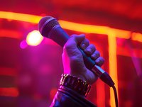 Hand holding microphone against vibrant red and pink stage lighting, capturing the energy of live performance at a concert or karaoke venue.