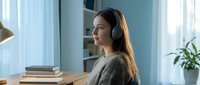 Young woman with long dark hair and East Asian ethnicity wearing wireless headphones while working at wooden desk in bright modern office space with blue walls, potted plant, and natural lighting.