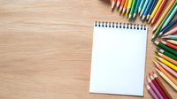 Blank spiral notepad with colorful pencils arranged on wooden desk background, copy space for text or artwork. Creative workspace setup for drawing and writing.
