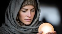 Young Caucasian woman in gray hooded scarf holding glowing orb with mystical light, fortune teller or spiritual healer concept for wellness design.