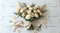 Elegant bridal bouquet with cream roses, eucalyptus and pampas grass on white wooden background with silk ribbon and scattered petals for wedding design.