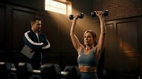 Athletic blonde woman lifting dumbbells overhead while male trainer observes in modern gym with brick walls and warm lighting for fitness motivation.