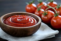 Fresh homemade tomato sauce in wooden bowl with swirl pattern and ripe cherry tomatoes on vine against dark background, close up view on natural linen napkin.