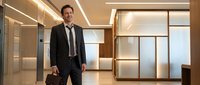 Confident male professional in formal business attire walking through modern office corridor with contemporary lighting and glass partitions, suitable for corporate and business concepts.