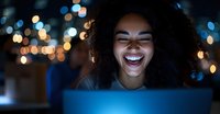 Young African American woman laughing while working on laptop at night, illuminated by screen glow and bokeh lights, expressing genuine joy and positive emotions.