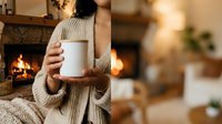 Woman in cozy beige sweater holding blank white mug by fireplace, warm autumn home interior with fire glowing in background, perfect for beverage branding and seasonal lifestyle marketing.