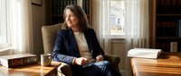 Professional middle-aged Caucasian businesswoman in navy suit sitting confidently in modern office chair by window, executive leadership concept.