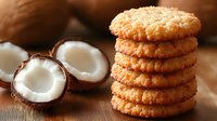 Stack of homemade coconut cookies with fresh coconut halves on wooden background, macro food photography showing crispy texture and golden brown color.