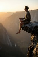 Young man meditating on cliff edge at sunset overlooking canyon valley. Peaceful mindfulness and spiritual wellness concept for mental health and relaxation.