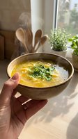 Steaming hot homemade soup with fresh herbs in ceramic bowl held by hand near sunny kitchen window with natural light and green plants in background.