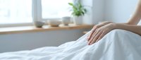 Young woman's hand gently resting on white bed linen in a bright, minimalist bedroom with a window and green plant in the background.