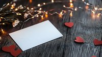 Blank white card with red paper hearts and glowing string lights on dark wooden background, romantic holiday mockup for Valentine message or love letter.