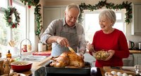 Senior Caucasian couple preparing holiday turkey dinner together in festive kitchen decorated with garland, sharing joyful cooking moment during celebration.