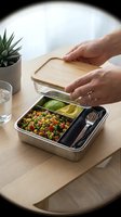 Person with medium skin tone opening wooden lid on compartmentalized lunch container filled with colorful vegetable salad, cucumber slice, and utensils on wooden table with succulent plant and water glass.