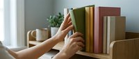 Woman selecting hardcover book from wooden shelf with various colored volumes and potted plant in bright interior space for reading, education, library, and home decor concepts.