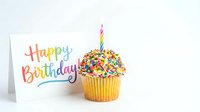 Colorful birthday cupcake with sprinkles and a lit candle beside a Happy Birthday greeting card on white background.