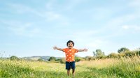 Young boy with curly dark hair and medium skin tone wearing bright orange shirt running through green meadow with arms outstretched, expressing joy and freedom outdoors.