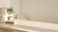 Minimalist home interior with natural sunlight streaming across white shelf displaying books, ceramic vase with olive branch and neutral decor elements.