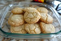 Fresh baked sugar cookies with crackled tops arranged in glass container on marble counter, showing classic homestyle texture and golden brown edges.