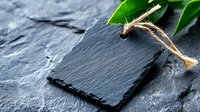Empty black slate stone tag with rope on dark background and green leaves, copy space for menu, price or product presentation.