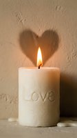 Burning white pillar candle with Love inscription casting heart-shaped shadow on beige wall, romantic Valentine atmosphere.