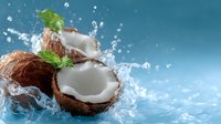 Fresh coconuts cracked open with splashing water drops and green mint leaves against blue background, dynamic food composition with white pulp and brown shell.