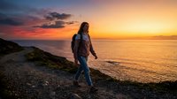 Young woman with long brown hair, wearing casual hiking attire and backpack, walking along rocky coastal cliff during golden hour sunset. Adventure travel and outdoor exploration concept.