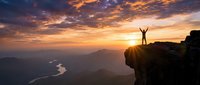 Person celebrating success on mountain cliff edge at golden sunrise with dramatic sky and valley river below for motivation concepts.