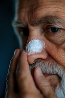 Close-up of senior Caucasian man applying moisturizing cream on nose, showing skincare routine and aging skin texture with white beard against dark background.