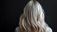 Platinum blonde wavy hair from behind on dark background shows modern hairstyling and coloring technique with soft natural waves and dimensional highlights.