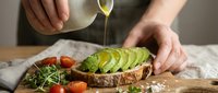 Hands drizzling olive oil over avocado toast with sliced avocado, cherry tomatoes and microgreens on rustic wooden cutting board for healthy breakfast.