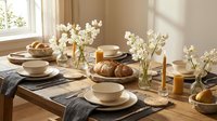 Elegant breakfast table setting with white flowers, ceramic dishes, fresh bread and orange juice in warm natural sunlight creating cozy morning atmosphere.