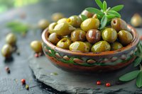 Fresh marinated green olives served in traditional ceramic bowl on black stone background with olive leaves and pink peppercorns for Mediterranean cuisine.