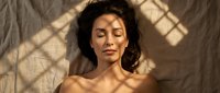 Peaceful young African American woman with curly hair relaxing with closed eyes in warm sunlight streaming through window creating geometric shadows on skin.