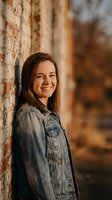 Young Caucasian woman in denim jacket smiling warmly while leaning against weathered brick wall during golden hour sunset for lifestyle photography.