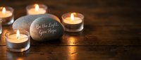 Inspirational stone with Be the Light Give Hope message surrounded by glowing tea light candles on rustic wooden surface creating warm spiritual atmosphere.