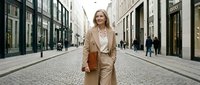 Confident middle-aged Caucasian businesswoman in beige coat walking on elegant European cobblestone shopping street holding leather folder.