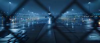 Moody urban nightscape through metal fence with rain reflections on wet asphalt, city lights creating atmospheric blue glow in foggy weather. Panoramic view.